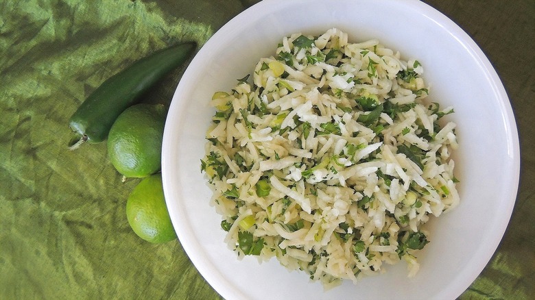 Top-down view of jicama coleslaw with green herbs and fresh lime in a white bowl