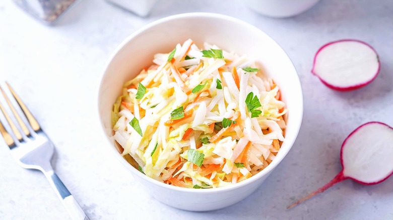 Coleslaw with cabbage, radish, and carrot in a white bowl next to a metal fork