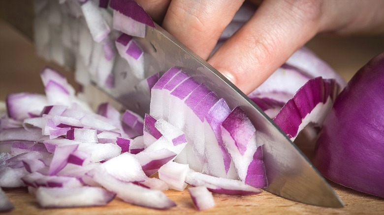 Hands chopping a red onion with a large knife on a wooden cutting board