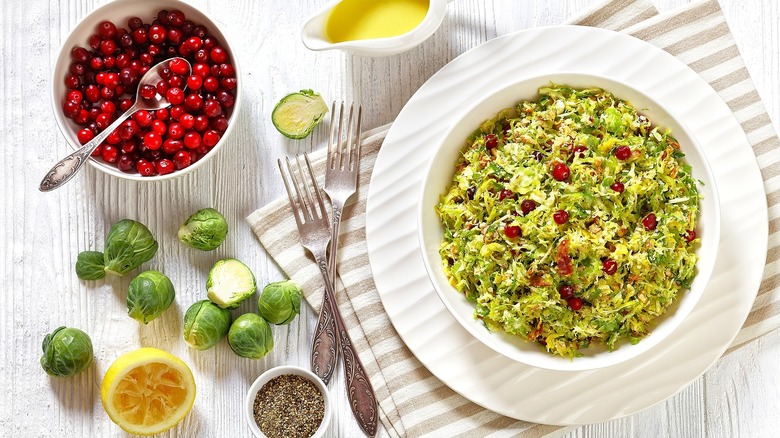 Top-down view of Brussels sprouts slaw with fresh cranberries in a white bowl on a white plate next to a bowl of whole cranberries