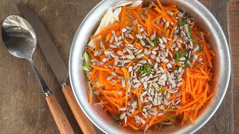 Top-down view of shredded carrots, cabbage, sunflower seeds, and green herbs in a silver bowl on a wooden surface