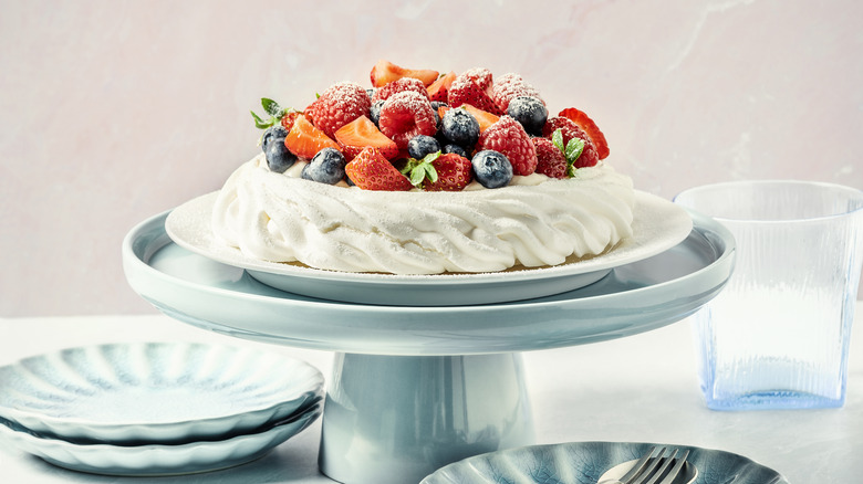 A pavlova on a blue cake stand topped with fresh fruit dusted with powdered sugar next to plates and glassware