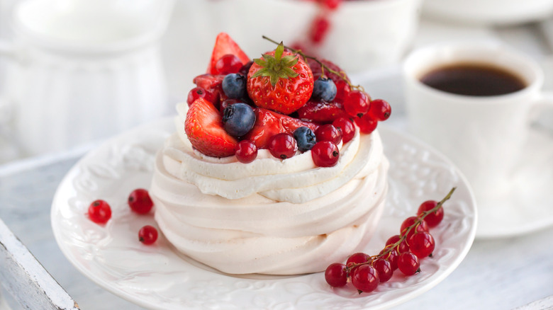 Image of a small pavlova topped with a variety of fresh berries in front of a mug of coffee