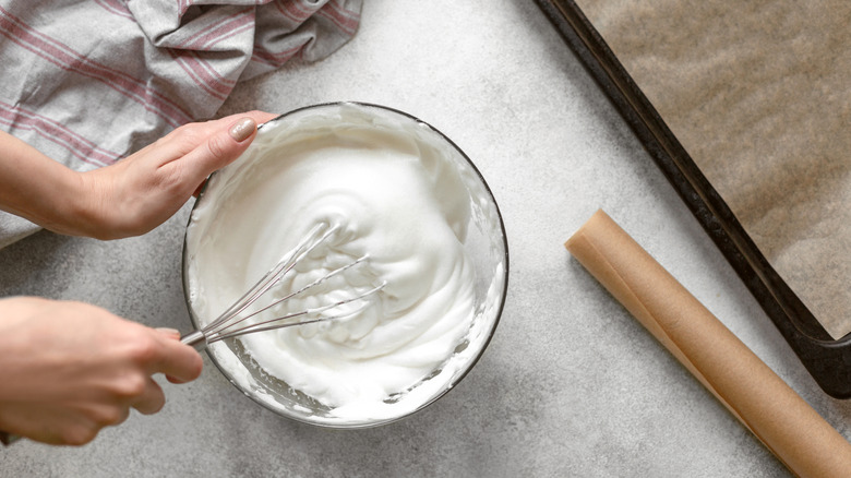 Making meringue by hand in a glass bowl with a metal whisk next to a lined sheet pan