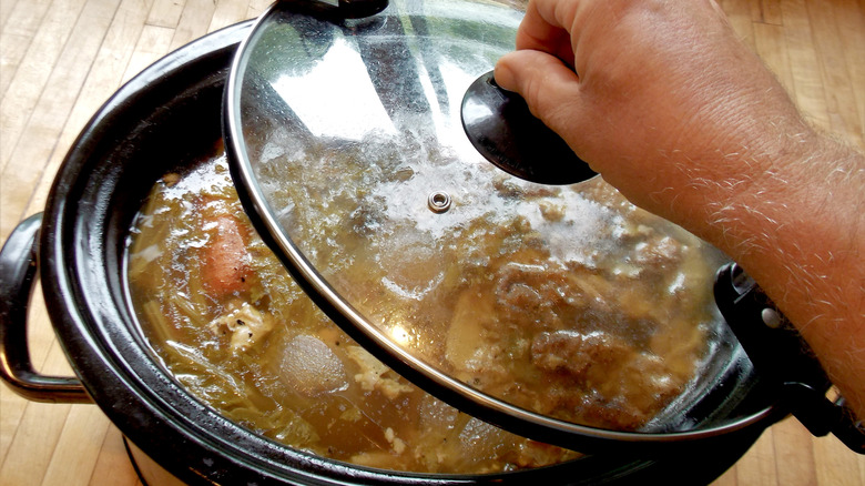 Person taking the lid off of a slow cooker filled with chicken soup.