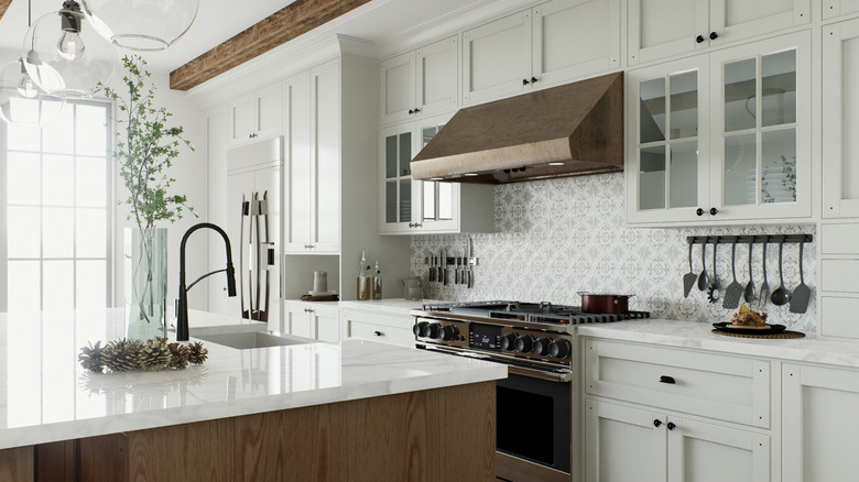 A mostly white kitchen with brown trim, featuring white marble countertops and bright light shining through French doors.