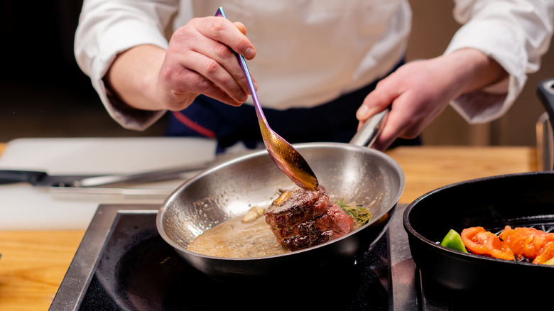 A chef spooning butter over steak in a stainless steel pan