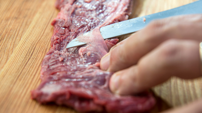 A long knife slices under a membrane on a raw skirt steak