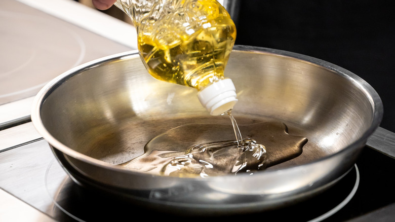 A bottle of cooking oil pours into a stainless steel pan on an electric stovetop