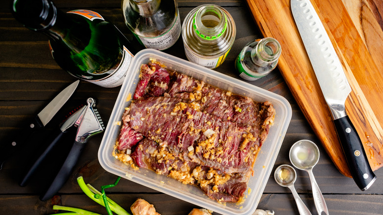 Skirt steak in a Tupperware container covered with ginger and garlic marinade, surrounded by open bottles and a wooden cutting board with a chef's knife