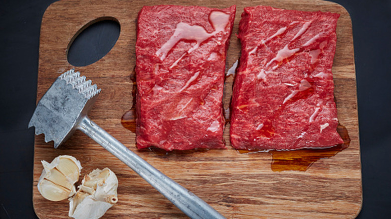 A wooden cutting board with two siirt steaks, a bulb of garlic, and a metal meat mallet