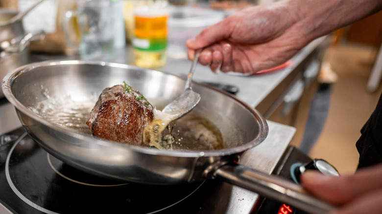 A person's hands are seen basting a steak in a tilted stainless steel pan.