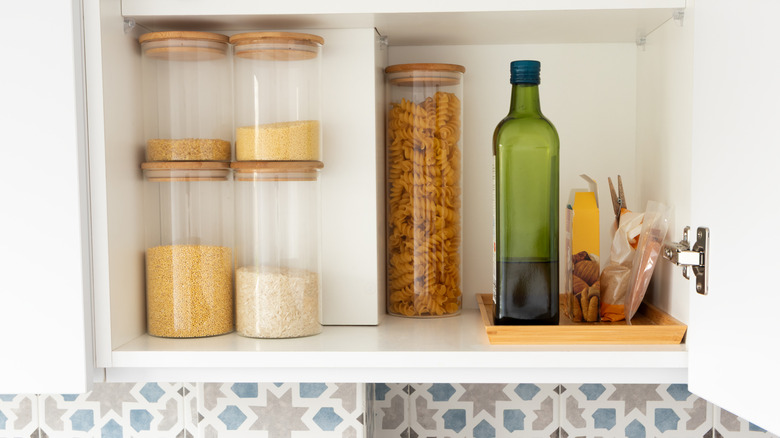 A green bottle of olive oil sits in a pantry next to a jar of pasta.