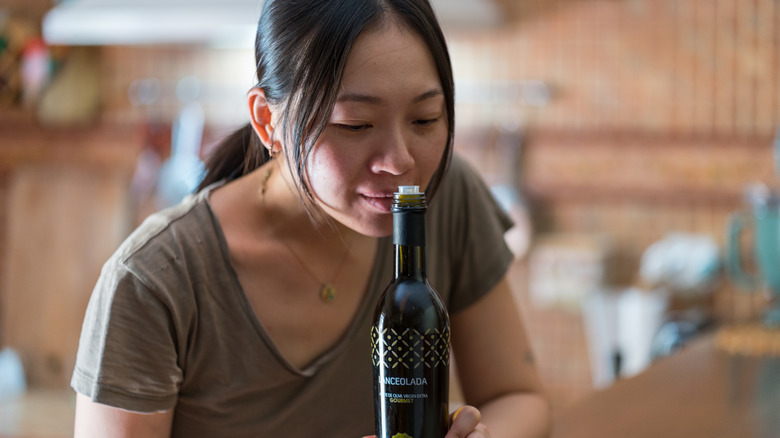 An Asian woman smells the top of an olive oil bottle.