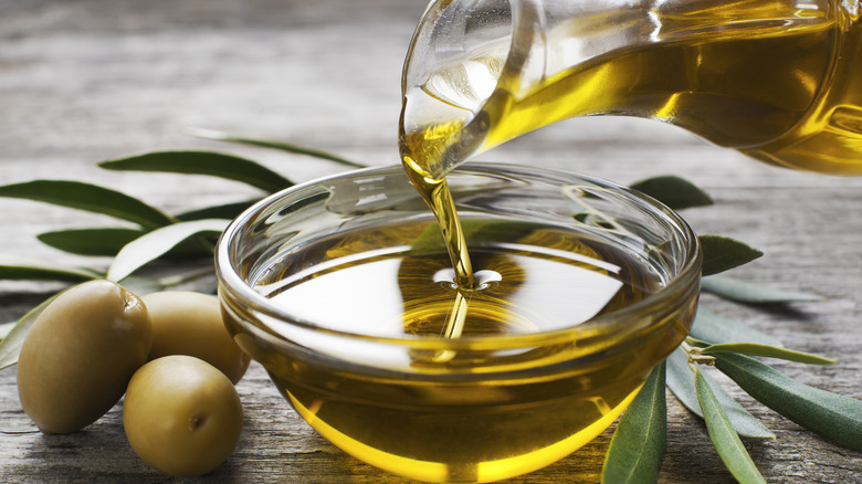 A jug of olive oil is poured into a smaller bowl with three olives next to it.