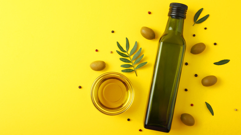 A green bottle of oil oil lays flat against a yellow background with olives and leaves.