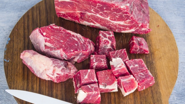 Raw beef in various stages of being cut, from roast to chunks, on cutting board with knife.