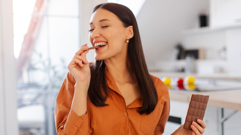 Happy woman with closed eyes enjoying taste of chocolate bar, sitting in kitchen at home