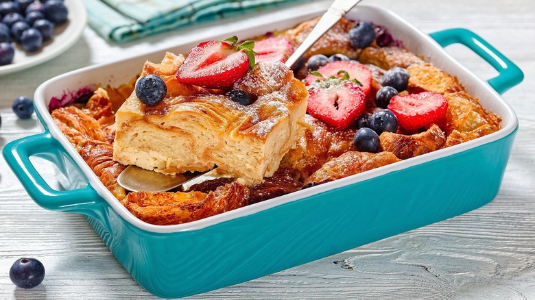 Croissant French toast casserole topped with powdered sugar, blueberries, and sliced strawberries in a turquoise baking dish