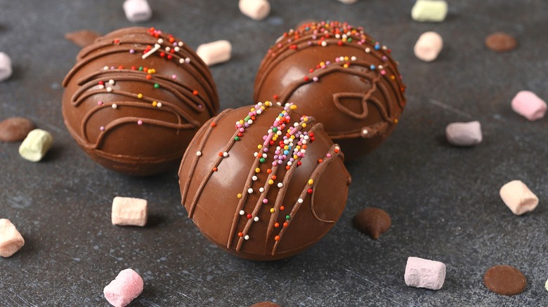 Three hot chocolate bombs with rainbow sprinkles surrounded by small pieces of chocolate and miniature marshmallows.