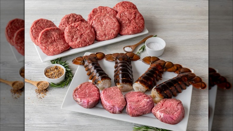 Selection of Angus beef and lobster tails on multiple white plates with seasonings on the side on countertop.