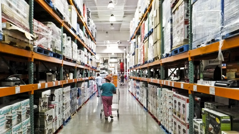 A shopper wearing a hat strolling down a Costco warehouse aisle stocked with different brands.