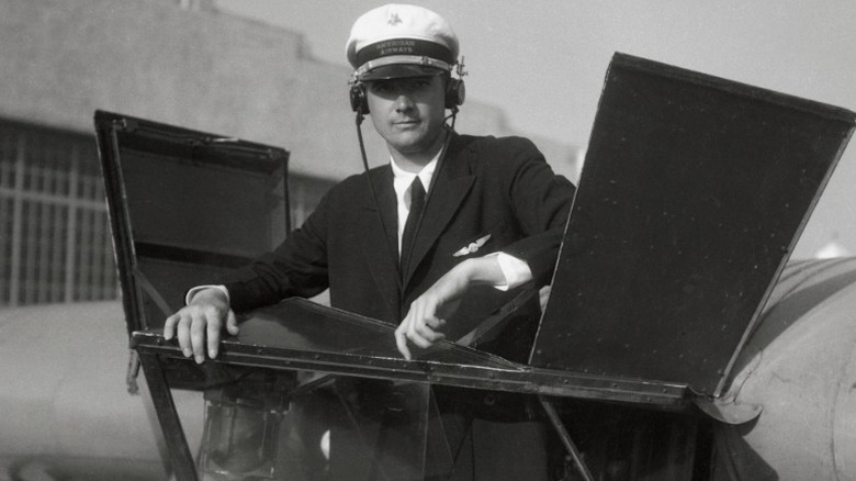 A young Howard Hughes wearing an airline pilot uniform with headphones on while standing on flight equipment outdoors