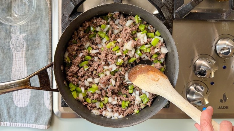 Stirring ground beef and chopped aromatic vegetables and herbs in skillet with wooden spoon