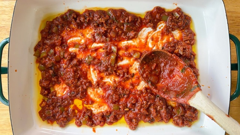 Using a wooden spoon to spread beef lasagna sauce in a 13 x 9-inch pan