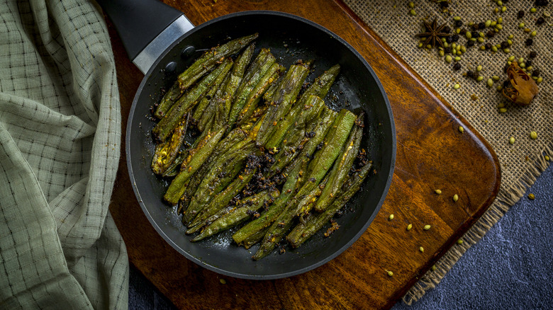 Top-down view of cooked okra in a metal skillet, on top of a cutting board and surrounded by whole spices