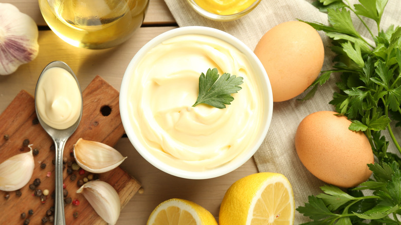 A bowl of homemade mayonnaise with a parsley leaf on top, surrounded by a spoon of mayonnaise, eggs in the shell, oil, garlic, peppercorns, and more parsley.
