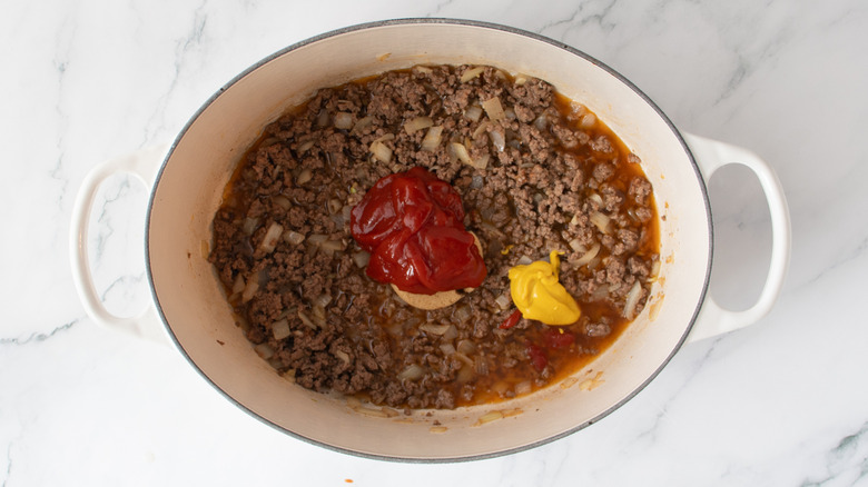 cooked ground beef and onions in a white pot with blobs of ketchup and mustard