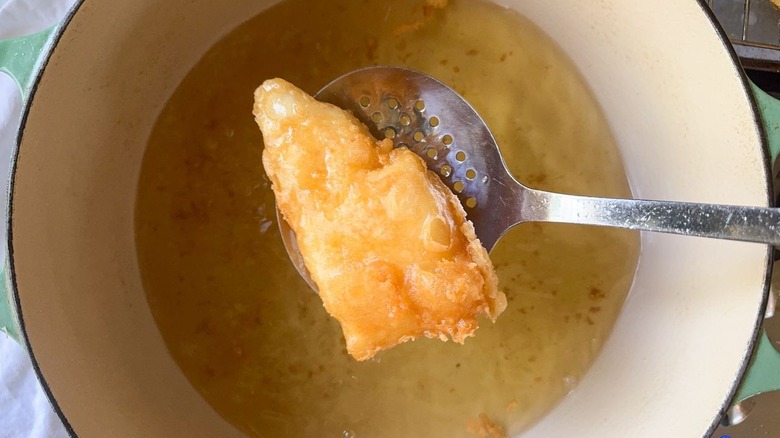 Beer-battered fried fish on slotted spoon above hot oil in pot on stove