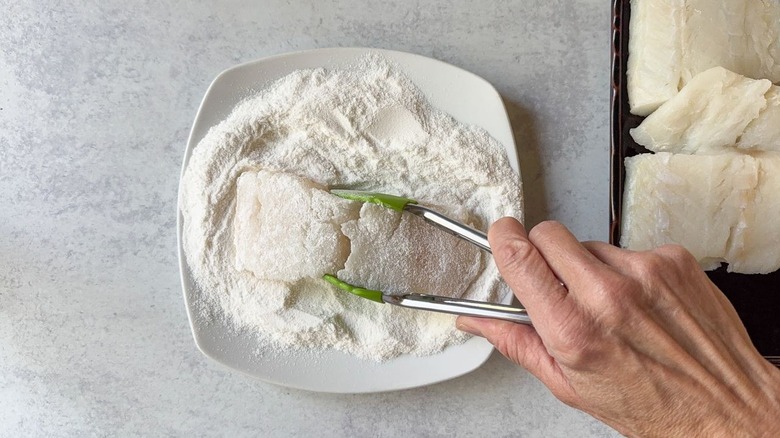 Dredging a cod filet in rice flour on a plate with tongs