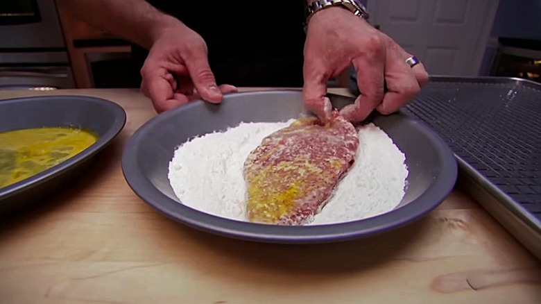 Alton Brown's hands dredging flattened steak into flour, egg wash, and then flour again before frying.