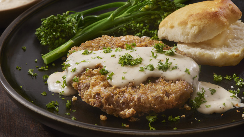 Chicken fried steak next to biscuit and broccolini on dark plate on table.