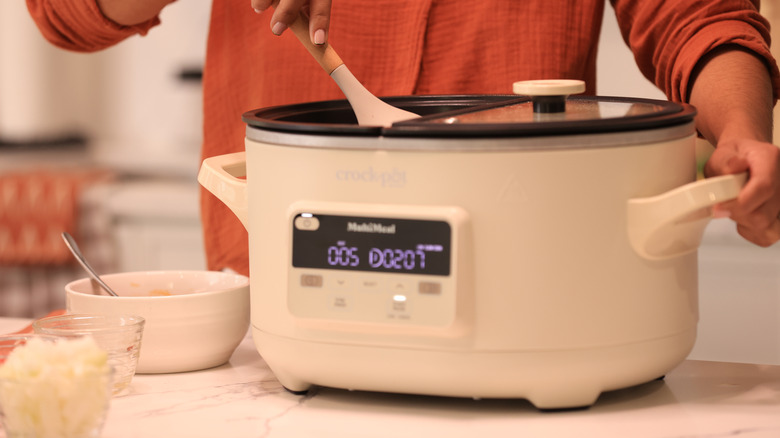 A person stirs the contents of a white Crock-Pot, dishes of ingredients to the left