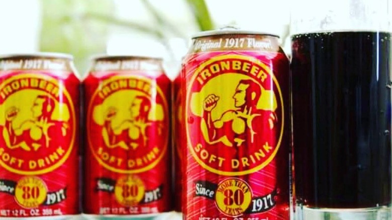 Three cans of Ironbeer alongside a glass of the dark soda