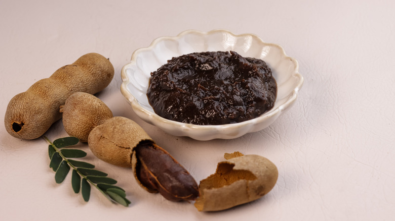A bowl of tamarind paste next to a whole and cut tamarind fruit on a white table