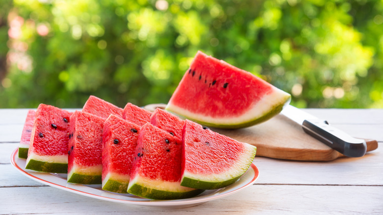 Watermelon triangles are arranged neatly on a plate with a watermelon wedge and a knife on a cutting board in the background.