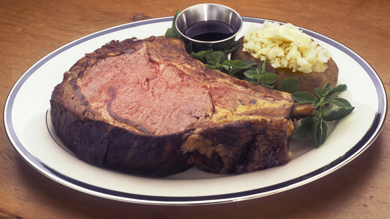 A large slice of cooked and tender prime rib on a plate with a baked potato and a side of beef jus