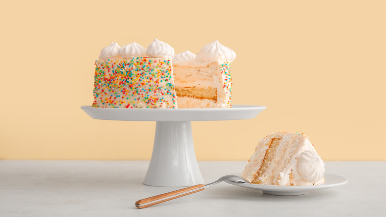 Layered birthday cake with sprinkles on a cake stand, with one slice on a plate