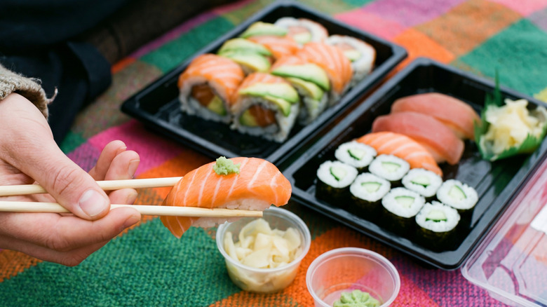 Salmon sushi in chopsticks with two trays of sushi in the background