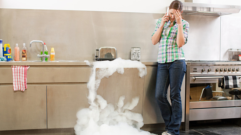 Stressed woman on the phone while watching a dishwasher overflow with soap suds
