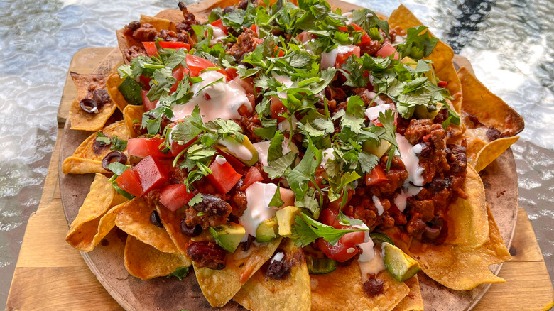 Loaded nachos piled high with chopped tomatoes, avocado, black olives, sour cream, and cilantro, served on a pizza stone.