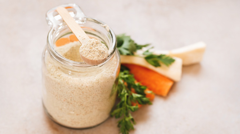 Glass mason jar of powdered soup mix with some in a wooden spoon balancing on top of the jar, with vegetables and herbs in the background