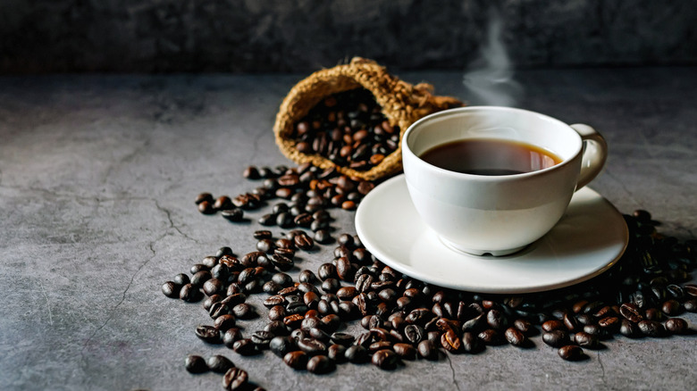 A steaming cup of fresh, hot, black coffee on a grey countertop, a bag of roasted coffee beans spill out behind it