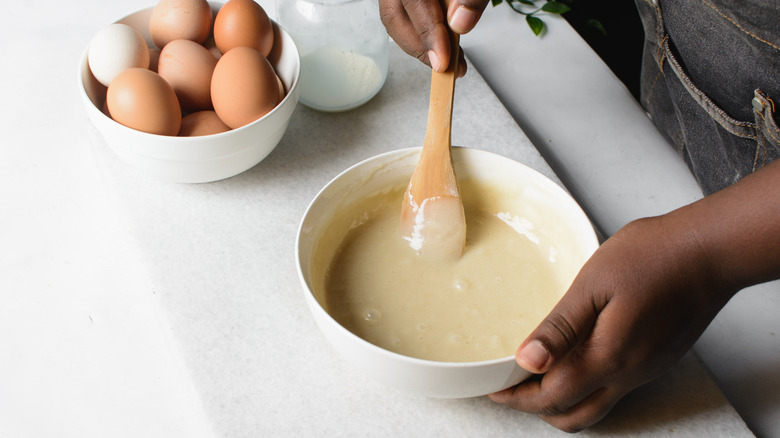 Close-up of a person's hands mixing cake batter in a bowl with a wooden spoon next to a bowl of eggs