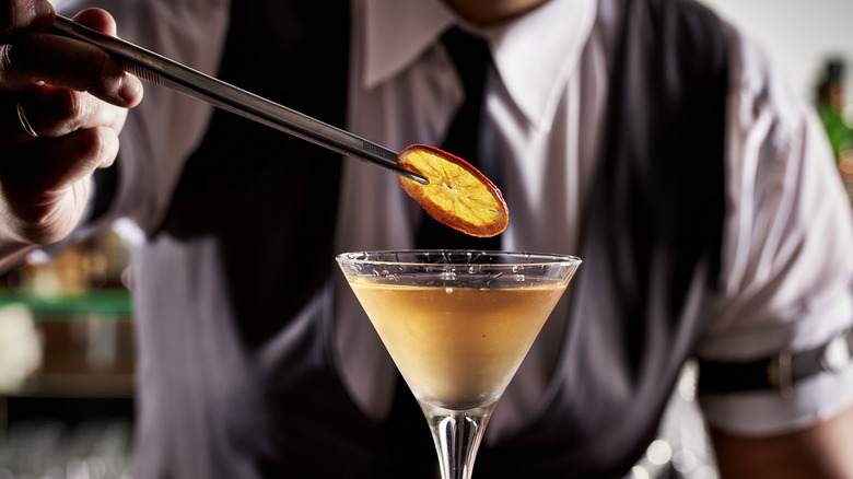 Bartender holding an orange slice over a cocktail in a martini glass.