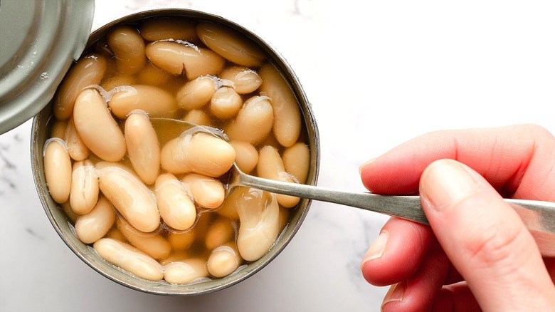 Hand holding a metal spoon in a can of white beans over a white marble surface